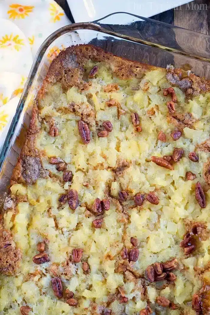 A close-up of a Gingerbread Dump Cake in a glass dish. The topping consists of chopped pecans and pieces of pineapple, creating a golden-brown crust. The edges are slightly crisp, and a patterned yellow and white towel is partially visible beside the dish.