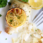 Air fryer fish fillet baked to perfection, topped with a lemon slice and garnished with parsley, served on a white plate with a fork.