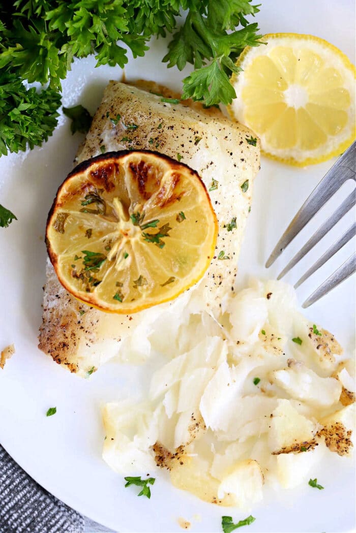 Air fryer fish fillet baked to perfection, topped with a lemon slice and garnished with parsley, served on a white plate with a fork.