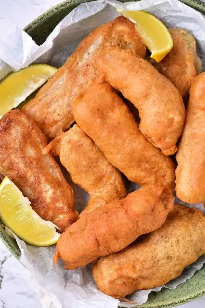 A plate of golden-brown beer-battered fish pieces is arranged on parchment paper in a bowl. Two lemon wedges are placed alongside the fish, adding a zesty contrast to the savory delight.