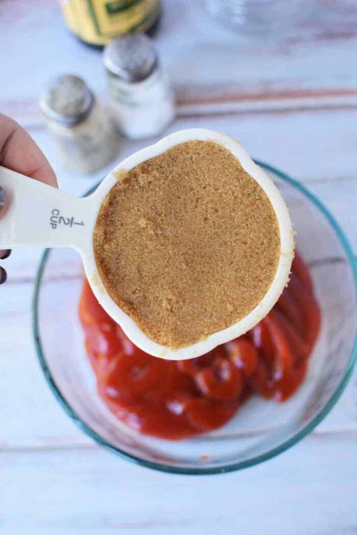 A hand holds a half-cup of brown sugar over a bowl of ketchup on a white table, showing the first step in How to Make Sweet Baby Rays Honey BBQ Sauce.