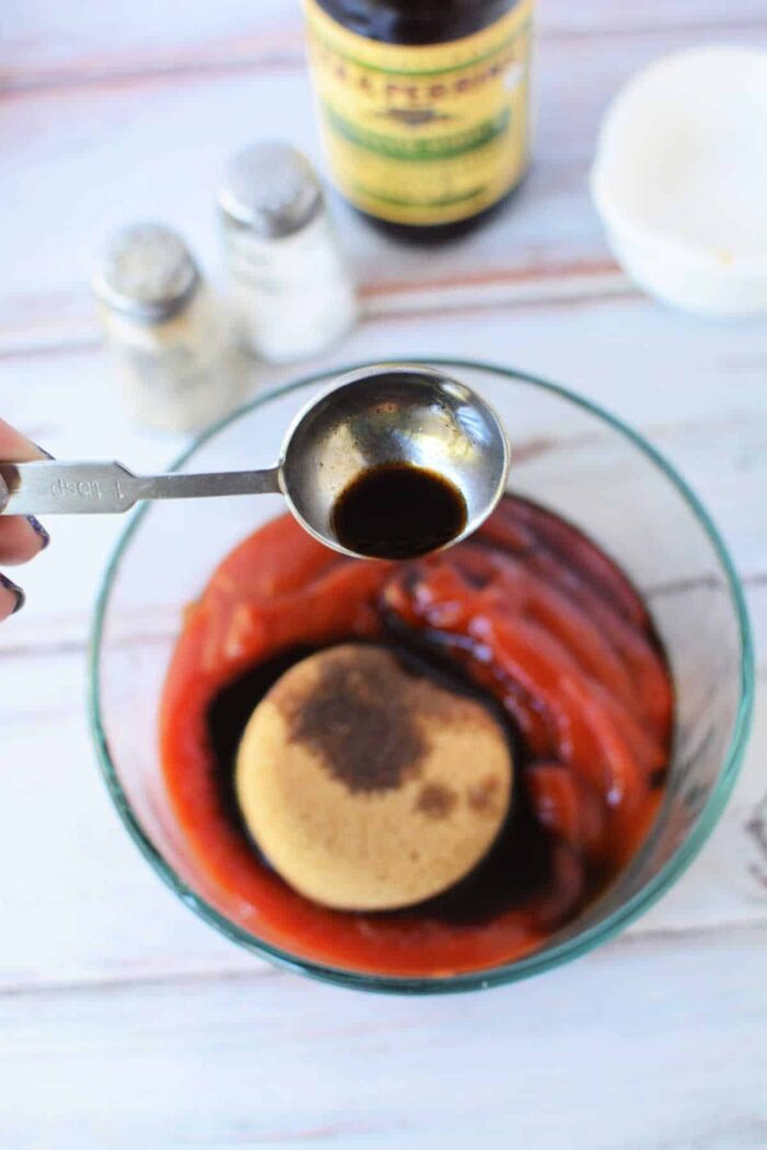 A spoon pours sauce into a bowl with ketchup, brown sugar, and spices on a white surface, illustrating How to Make Sweet Baby Rays Honey BBQ Sauce at home.