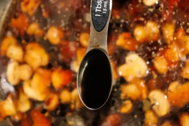 Close-up of a tablespoon labeled with 1 Tbsp, 15 mL, hovering above a pot of chickpeas, tomatoes, and spices. The spoon holds brown gravy, likely a rich seasoning for a meatloaf recipe.