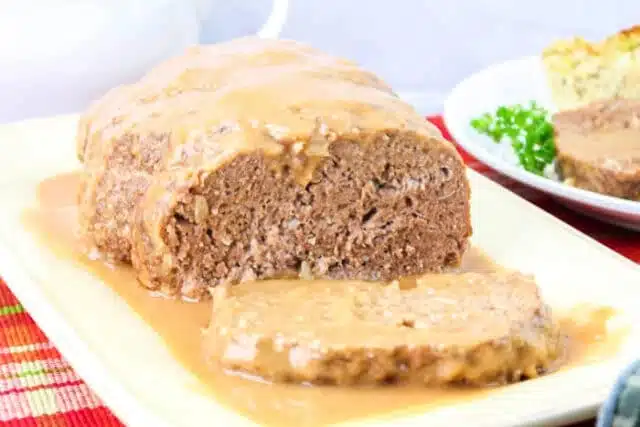 A plate of meatloaf topped with rich brown gravy, with one slice cut from the loaf. A partial view of a second plate featuring another slice and a garnish of parsley peeks through, all set on a multicolored placemat, perfectly showcasing this savory meatloaf recipe.