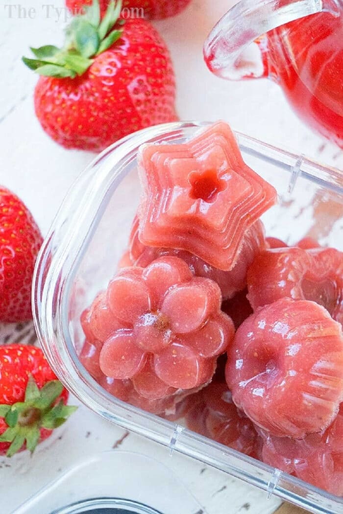 If you've never made homemade fruit snacks it's easier than you think! Only 4 ingredients to make sweet jellies complete with Vitamin C and real fruit! #homemade #fruitsnacks #jellies #strawberry #snacks Pink gelatin candies shaped like a star, flower, and bundt, with fresh strawberries nearby—perfect homemade fruit snacks for a delightful treat.