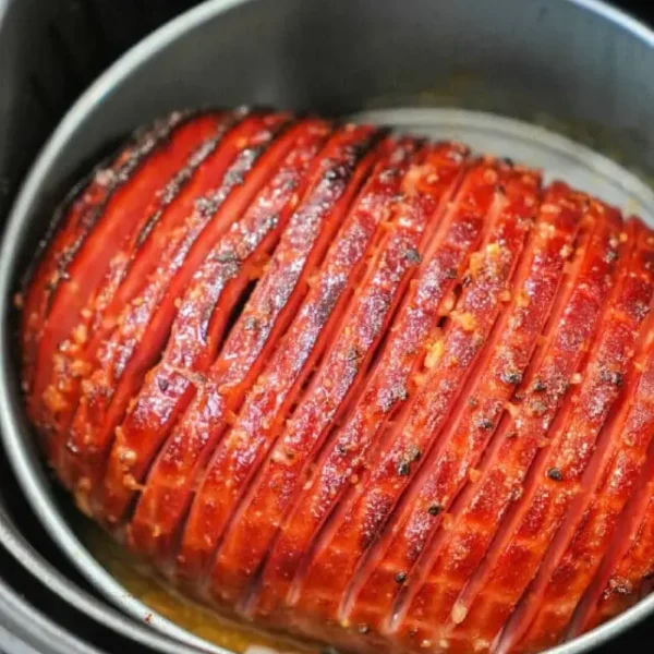 A meatloaf with a neatly sliced top layer, seasoned to perfection, is cooking alongside slices of ham in an air fryer. The meatloaf's slightly caramelized exterior suggests it's almost fully cooked.