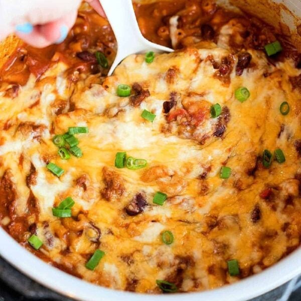 A hand holding a white spoon is stirring a cheesy, baked pressure cooker chili mac topped with green onions in a pot. The dish appears to have a tomato-based sauce and melted cheese.