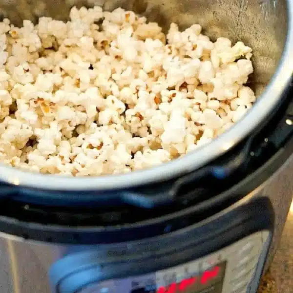 The Instant Pot is filled with a mountain of popcorn, its gleaming metallic interior adding a modern twist to this classic snack. The digital control panel stands out, while the brown and tan speckled countertop beneath exudes a cozy charm.