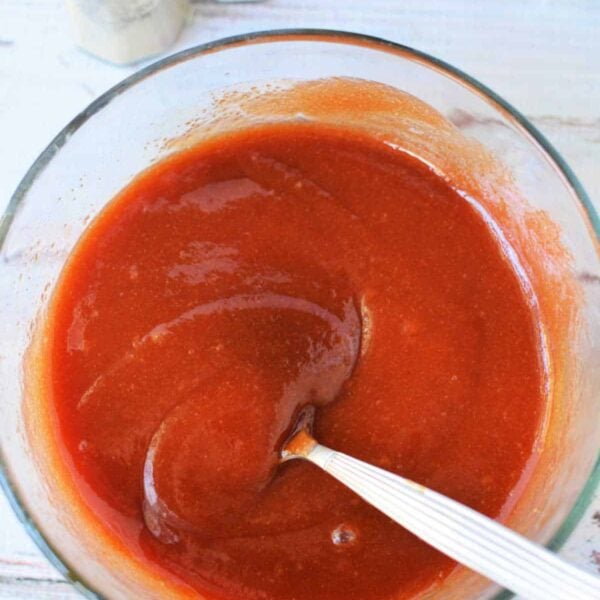 A glass bowl of reddish-brown sauce with a spoon, next to salt and pepper shakers on a white surface, perfect for those curious about how to make Sweet Baby Rays Honey BBQ Sauce at home.