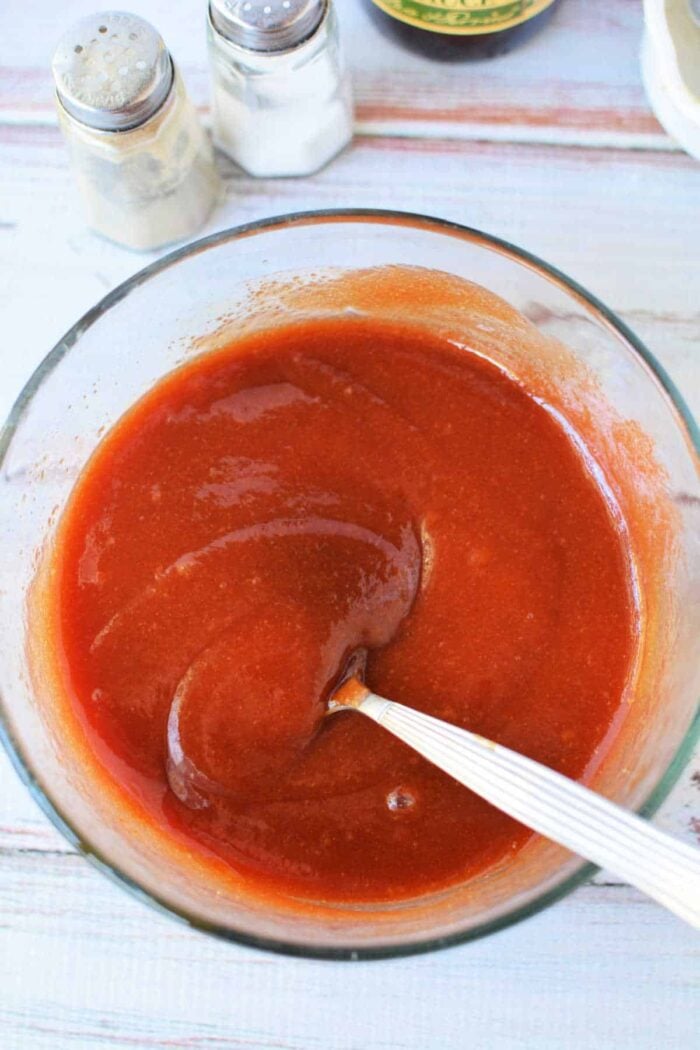 A glass bowl of reddish-brown sauce with a spoon, next to salt and pepper shakers on a white surface, perfect for those curious about how to make Sweet Baby Rays Honey BBQ Sauce at home.