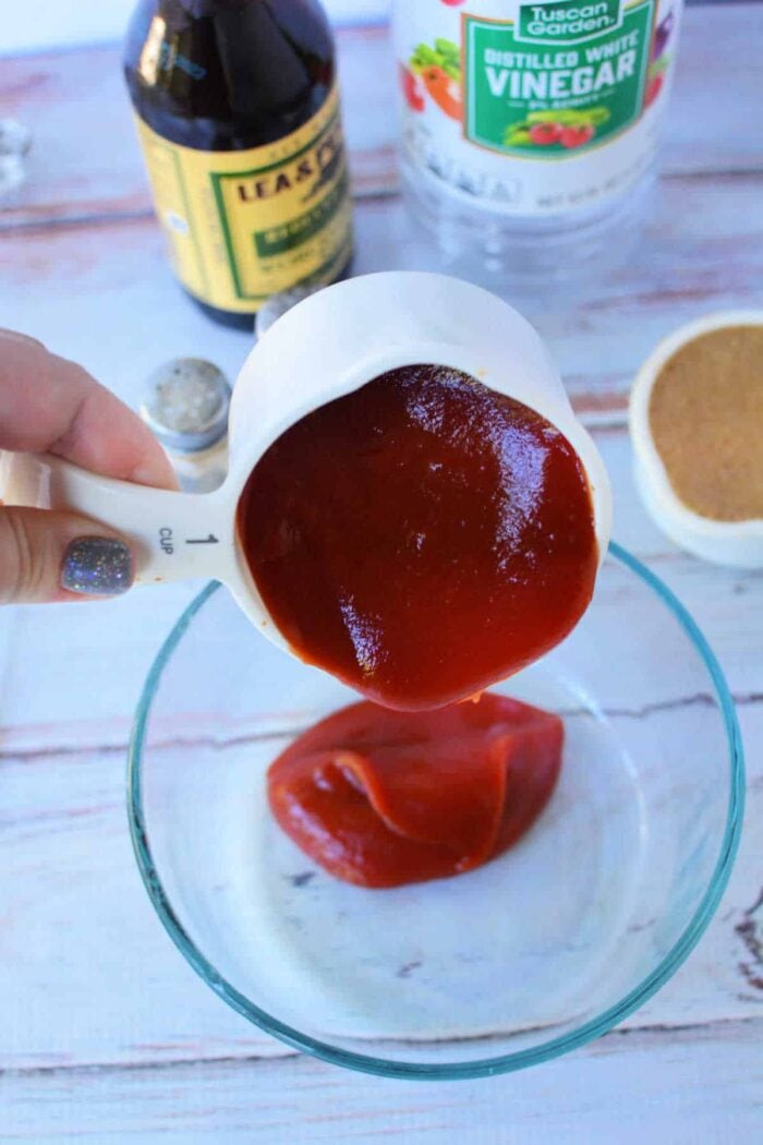 A hand pours ketchup from a measuring cup into a glass bowl, with vinegar and Worcestershire sauce nearby—key steps in learning How to Make Sweet Baby Rays Honey BBQ Sauce.
