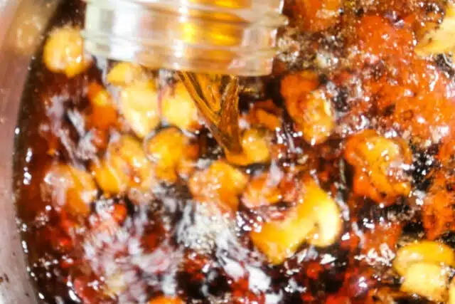 A jar is pouring a brown gravy over a mixture of nuts and seeds in a pan. The contents appear to be simmering, with a variety of textures and colors blending, suggesting a preparation process for either a sweet or savory dish, reminiscent of ingredients in meatloaf recipes.