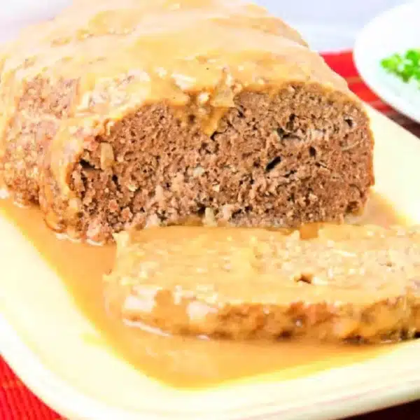 A loaf of meatloaf drenched in rich brown gravy for meatloaf recipe perfection sits on a light-colored platter. One slice is cut in the foreground. The dish rests on a red-and-yellow striped cloth, accompanied by a small bowl of green garnish in the background.