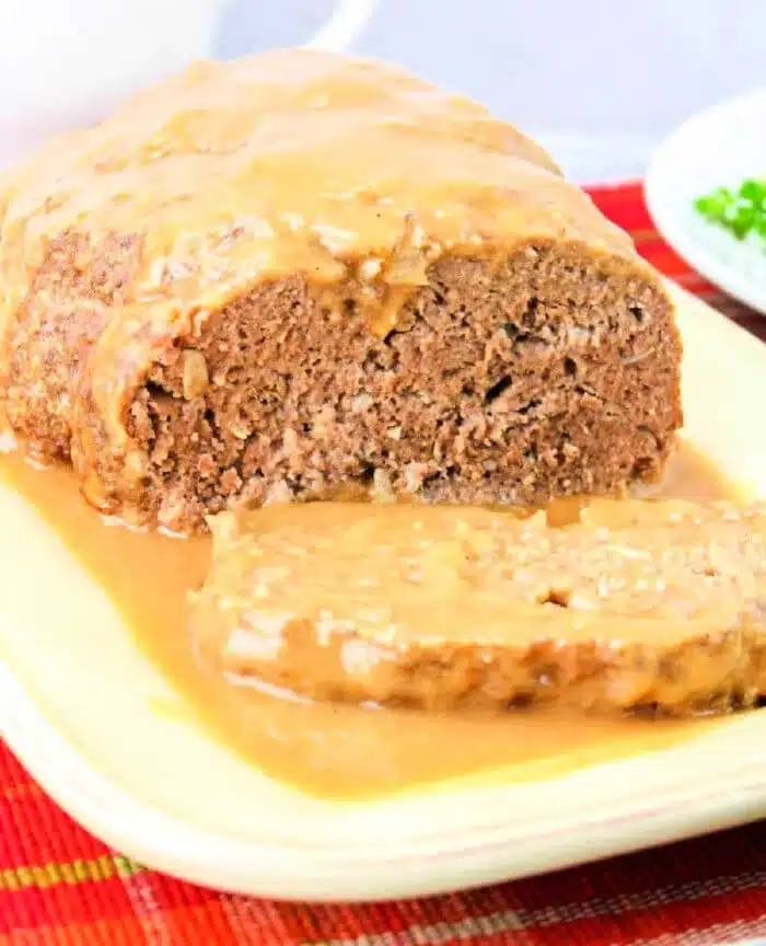 A loaf of meatloaf drenched in rich brown gravy for meatloaf recipe perfection sits on a light-colored platter. One slice is cut in the foreground. The dish rests on a red-and-yellow striped cloth, accompanied by a small bowl of green garnish in the background.