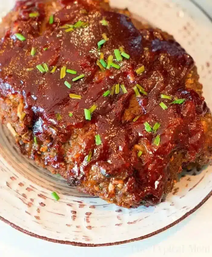 A plated portion of Ninja Foodi meatloaf topped with a glossy layer of barbecue sauce and garnished with chopped chives. The meatloaf sits on a white ceramic plate with dark streak patterns.