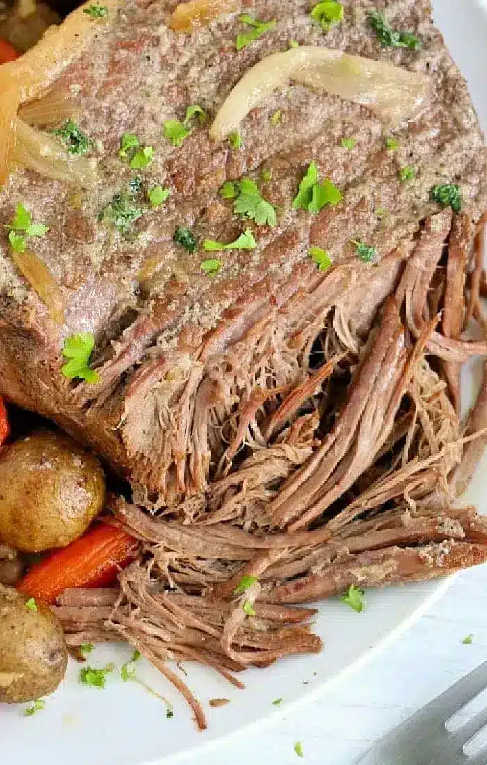 A close-up of an air fryer roast beef with shredded meat, garnished with chopped parsley. Potatoes and carrots are partially visible beneath the roast, which has a layer of cooked onions on top. The dish is presented on a white plate.