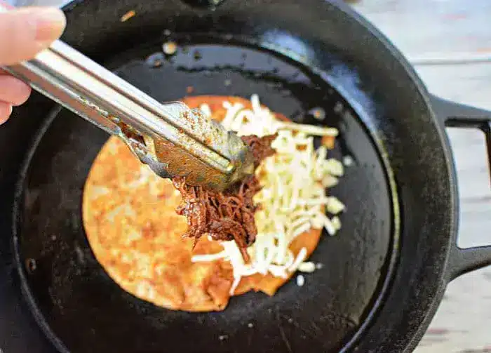 A hand holds metal tongs, placing shredded beef onto a tortilla with melted cheese in a black skillet. The tortilla appears partially folded, suggesting the preparation of a quesadilla, while a drizzle of savory birria tacos sauce enhances its flavor.