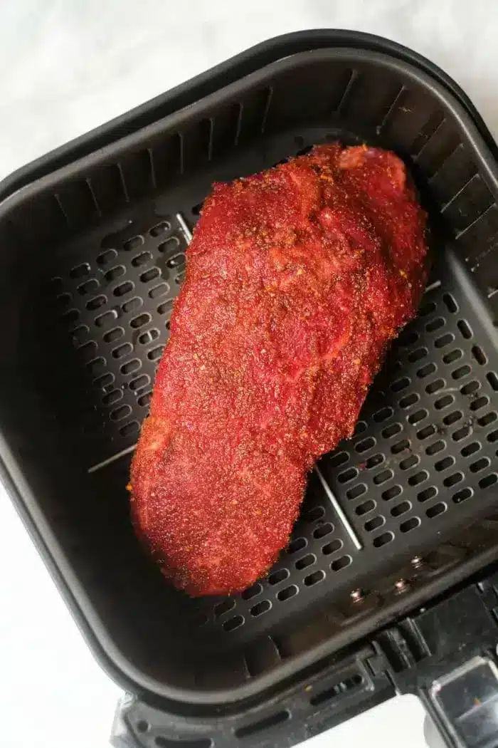 A piece of seasoned roast beef is placed in an air fryer basket. The meat has a reddish-brown crust, suggesting it has been coated with a spice rub. The air fryer basket has a dark non-stick surface with perforations for optimal air circulation.