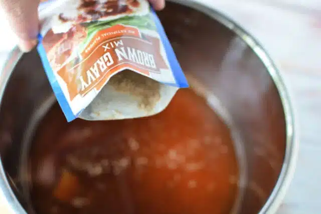 A hand is pouring a packet of brown gravy for meatloaf recipe mix into a metal pot filled with liquid. The contents are partially visible as they blend into the pot.