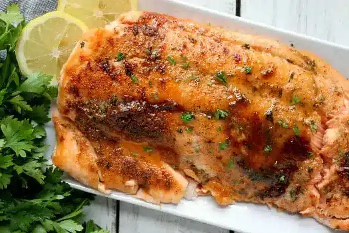 A rectangular white plate holds a cooked piece of glazed salmon, crisped to perfection in an air fryer and garnished with chopped herbs. The salmon is surrounded by sprigs of fresh parsley and two lemon slices. The plate is positioned on a white wooden surface.