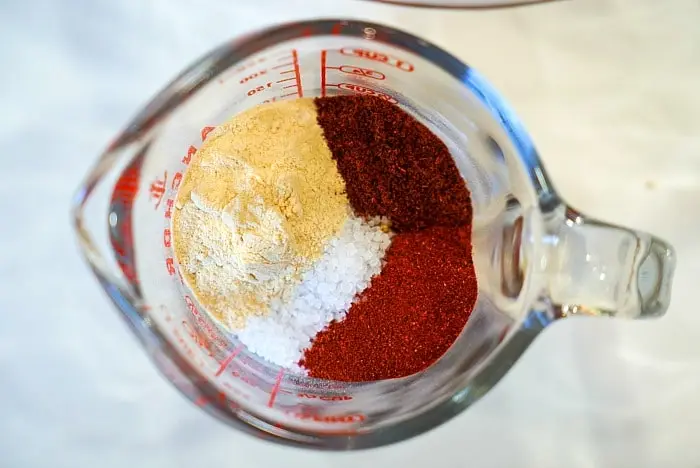 A glass measuring cup viewed from the top, filled with four different spices in partial portions. The spices include a light beige powder, a coarse white powder, and two different shades of red powder, each distinctly separated within the measuring cup—perfect for seasoning your next air fryer roast beef.