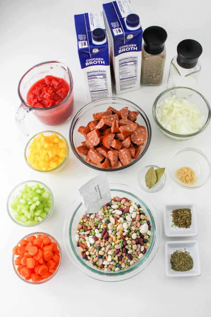 Assorted 15 Bean Soup Crock Pot ingredients in bowls: beans, sausage, vegetables, canned tomatoes, broth, and spices on a white background.