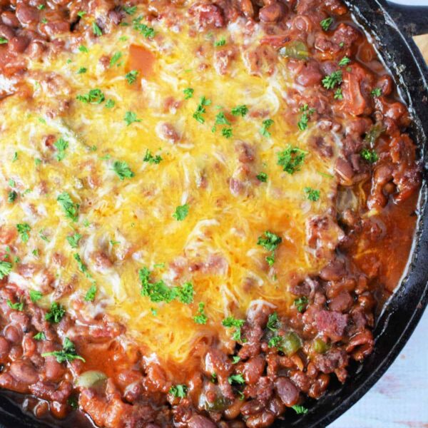 A cast iron skillet filled with baked bean casserole topped with a layer of melted cheese and garnished with chopped parsley. The dish is placed on a light wooden surface.