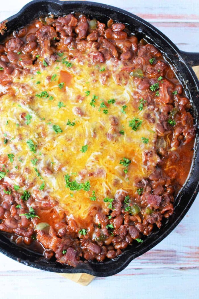 A cast iron skillet filled with baked bean casserole topped with a layer of melted cheese and garnished with chopped parsley. The dish is placed on a light wooden surface.