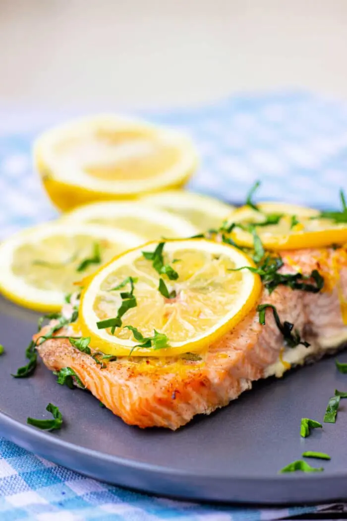 A piece of baked salmon garnished with lemon slices and chopped herbs is on a dark plate. 