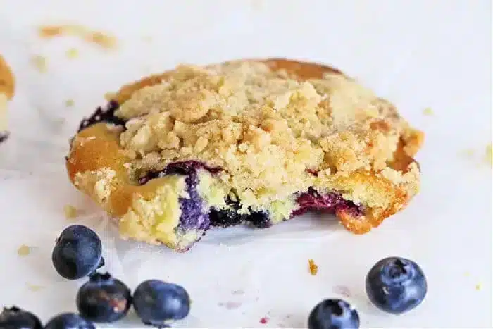 A crumbly pastry reminiscent of blueberry muffin tops, with a rich blueberry filling and topped with streusel, sits on a white surface. Several whole blueberries are scattered around.