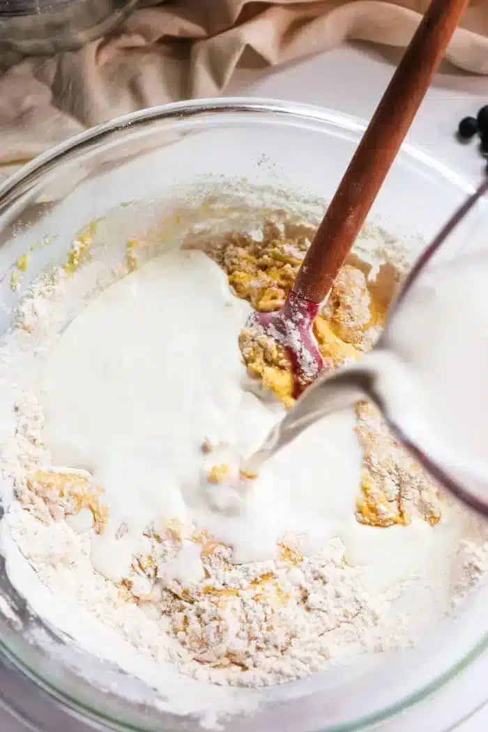 A glass mixing bowl contains flour and egg batter being mixed with a wooden spoon. A pitcher pours liquid, likely milk, into the bowl, perfect for making blueberry muffin tops. The background shows part of a beige cloth and some blueberries on the table.