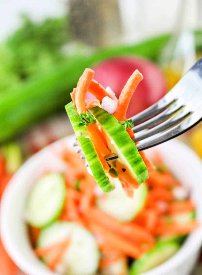 A fork holds a bite-sized portion of Carrot and Cucumber Salad, featuring cucumber slices, carrot strips, and chopped red onions. In the background, there is a bowl of similar salad ingredients and some vegetables.