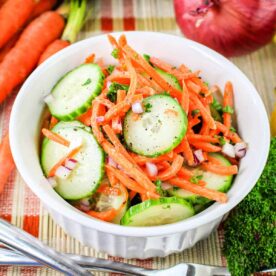 A white bowl filled with a Carrot and Cucumber Salad, featuring sliced cucumbers, julienned carrots, and chopped red onions, garnished with herbs. The bowl sits on a woven mat, surrounded by whole carrots, a cucumber, a red onion, and parsley. A fork and knife rest beside the bowl.