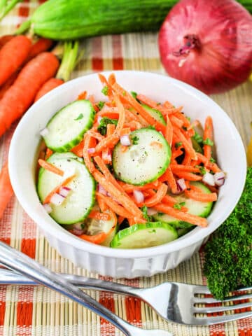 A white bowl filled with a Carrot and Cucumber Salad, featuring sliced cucumbers, julienned carrots, and chopped red onions, garnished with herbs. The bowl sits on a woven mat, surrounded by whole carrots, a cucumber, a red onion, and parsley. A fork and knife rest beside the bowl.