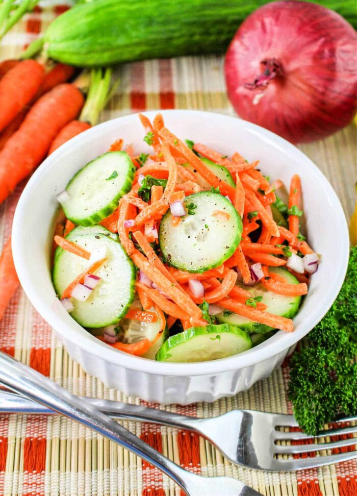 A white bowl filled with a Carrot and Cucumber Salad, featuring sliced cucumbers, julienned carrots, and chopped red onions, garnished with herbs. The bowl sits on a woven mat, surrounded by whole carrots, a cucumber, a red onion, and parsley. A fork and knife rest beside the bowl.