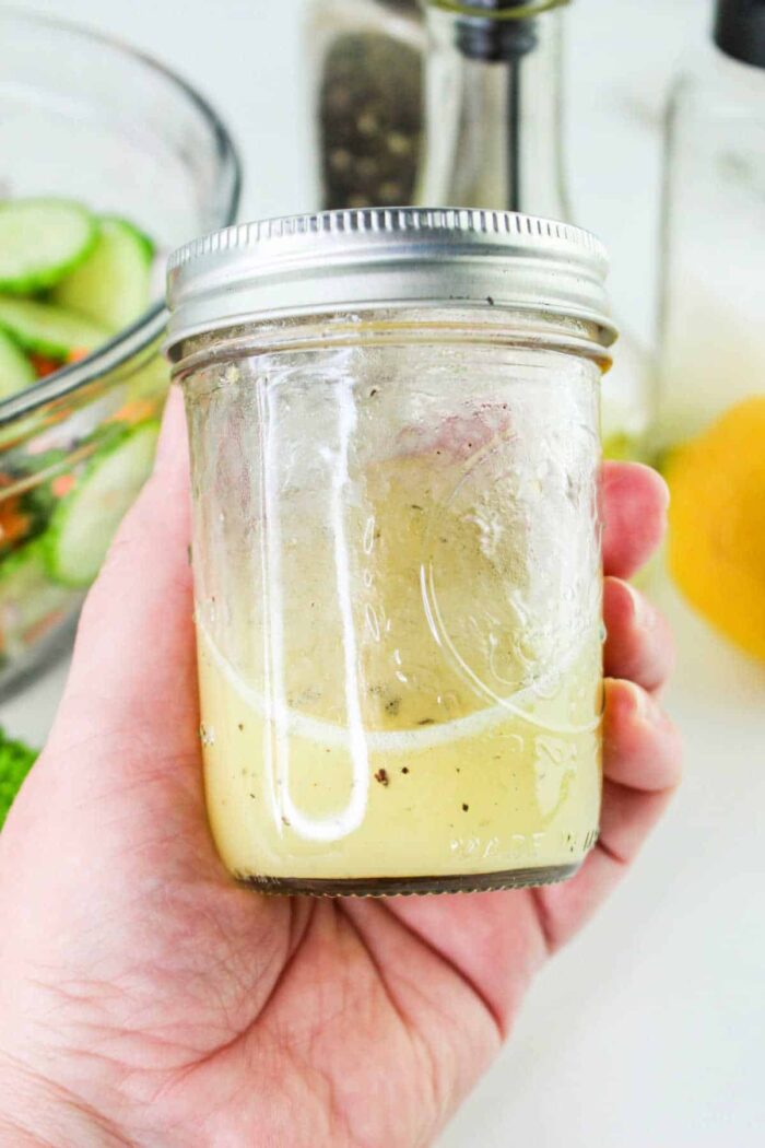 A hand is holding a glass jar filled halfway with light-colored salad dressing, perfect for a refreshing Carrot and Cucumber Salad. The jar has a metal screw-top lid. In the background, there is a bowl of sliced cucumbers, a pepper grinder, a bottle, and a lemon.