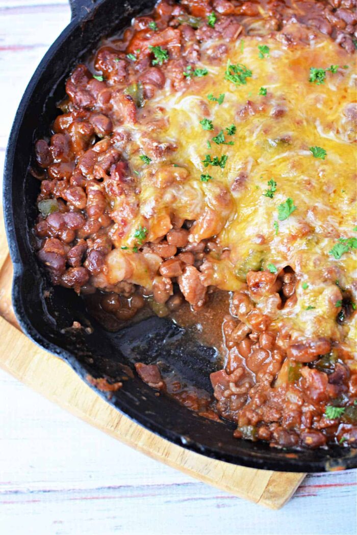 A cast-iron skillet filled with baked bean casserole topped with melted cheese and garnished with chopped parsley sits on a wooden trivet. The dish appears to be freshly cooked, with a portion already served from the skillet.