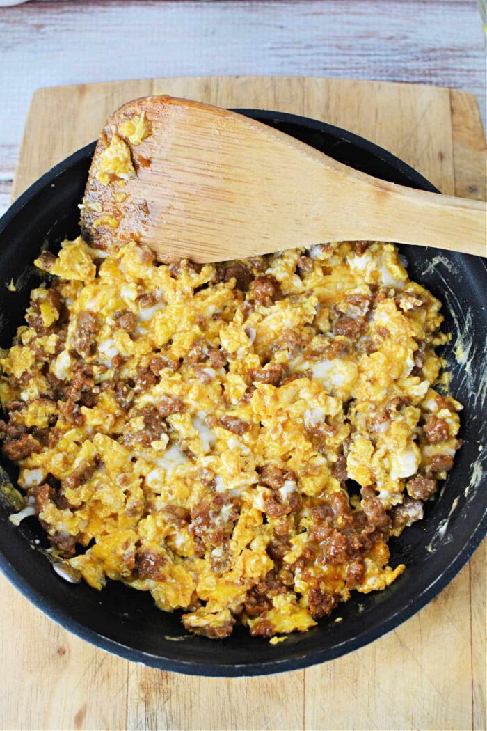 A frying pan on a wooden cutting board contains scrambled eggs mixed with ground sausage. A wooden spatula rests on the edge of the pan, partially covered in the egg and sausage mixture—a perfect demonstration of how to cook chorizo and eggs.