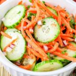 A fresh Carrot and Cucumber Salad in a white bowl contains thin slices of cucumber, grated carrots, and finely chopped red onion, garnished with chopped parsley. The bowl is on a woven mat with a fork and spoon nearby, and vegetables can be seen in the background.