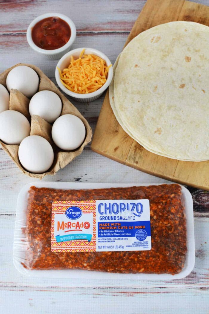 A wooden surface displays tortillas on a cutting board, eggs in a carton, grated cheddar cheese in a bowl, salsa in a small bowl, and a package of Kroger Mercado ground chorizo sausage—perfect ingredients for learning how to cook chorizo and eggs.