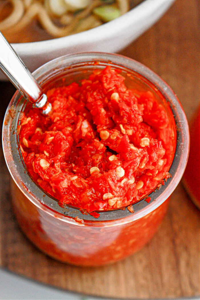 A clear glass jar filled with chunky garlic chili paste, showing visible seeds and a metal spoon inserted into the jar. It sits on a wooden surface with part of a white bowl containing food visible in the background.