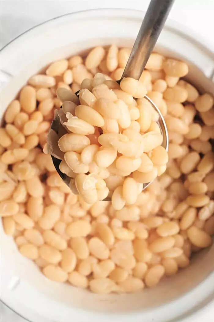 A ladle holding cooked navy beans is being lifted from a pot filled with more cooked beans. The beans are light beige in color and appear soft and tender, similar to how to cook Great Northern beans. The ladle is metal with a long handle.