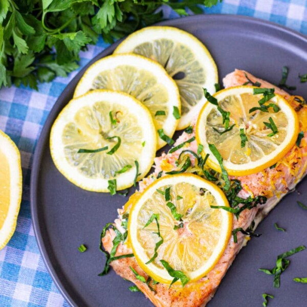 A cooked salmon fillet topped with lemon slices and chopped herbs is served on a gray plate, sitting on a blue and white checkered tablecloth. Fresh parsley and additional lemon slices are placed around the plate. Wondering how long to bake salmon at 350? This dish is perfectly timed for deliciousness.