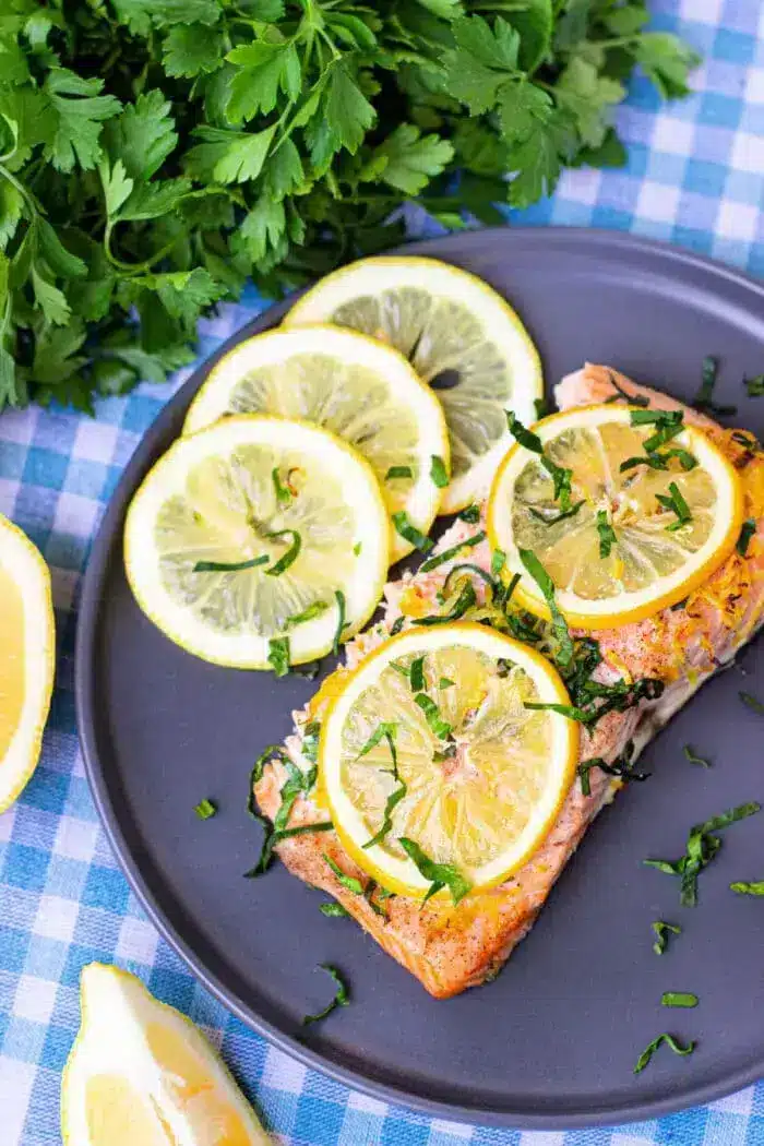 A plate containing a piece of cooked salmon topped with lemon slices and chopped green herbs. 