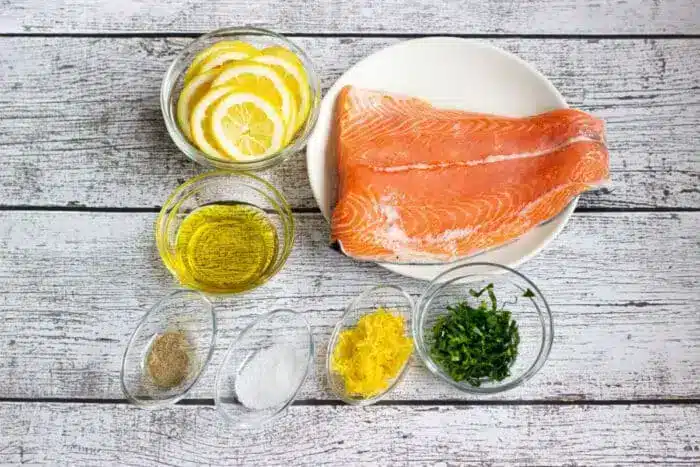 A plate with a raw salmon fillets is surrounded by small bowls containing lemon slices, olive oil, black pepper, salt, lemon zest, and chopped herbs, all placed on a white wooden surface. 