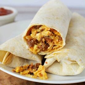 A white plate holds two burritos made with flour tortillas. One burrito is partially cut open, revealing a filling of scrambled eggs, sausage, and melted cheese. The scene offers a tempting glimpse into how to cook chorizo and eggs. Brown sauce in a small white bowl appears blurred in the background.