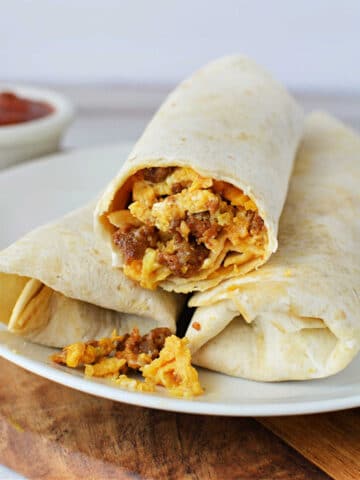 A white plate holds two burritos made with flour tortillas. One burrito is partially cut open, revealing a filling of scrambled eggs, sausage, and melted cheese. The scene offers a tempting glimpse into how to cook chorizo and eggs. Brown sauce in a small white bowl appears blurred in the background.