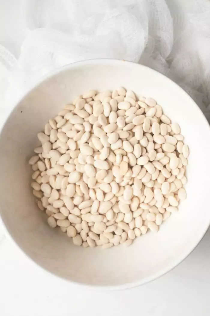 A white bowl filled with dried Great Northern beans is placed on a light-colored surface. 