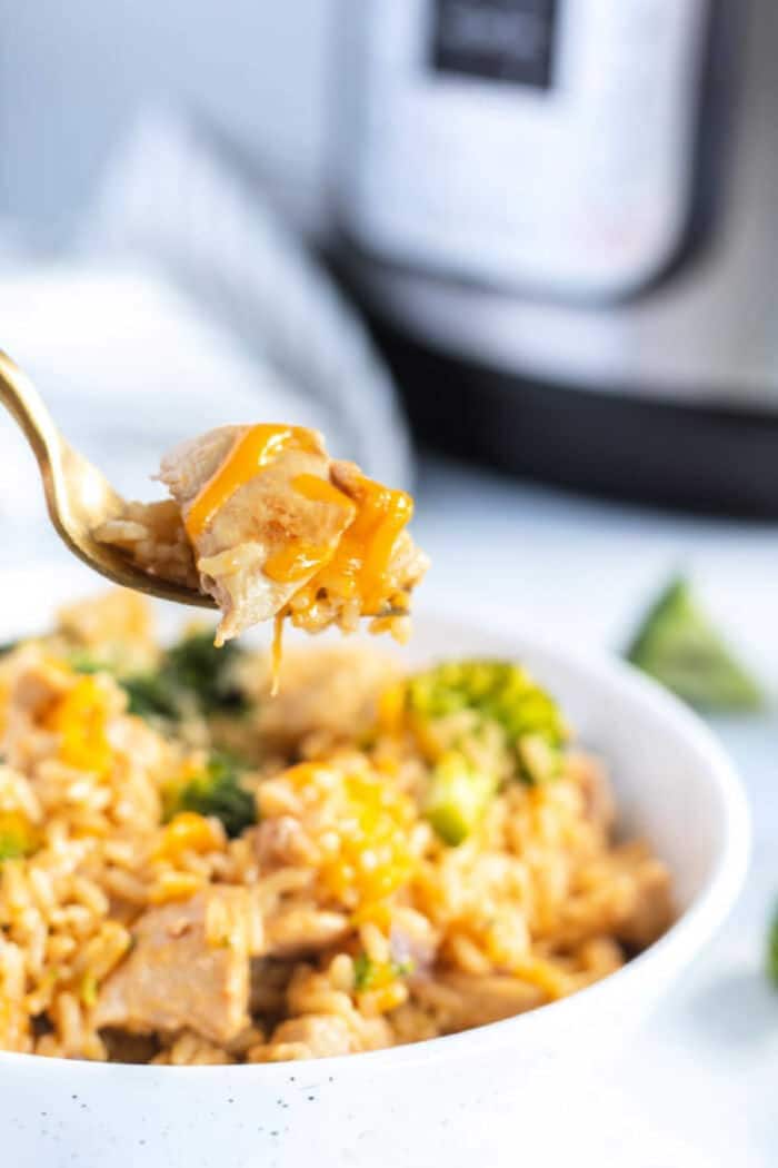 A fork holds up a bite of instant pot cheesy chicken and rice above a bowl brimming with the same comforting dish, with an Instant Pot in the background.