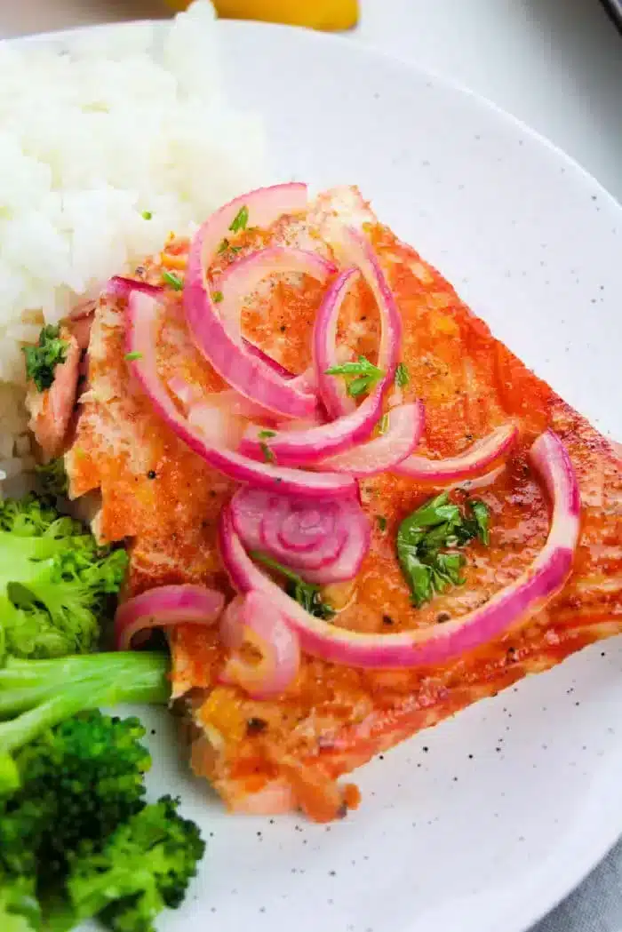 A close-up of a plate featuring a piece of succulent salmon topped with sliced red onions and herbs. The plate also includes servings of white rice and steamed broccoli.